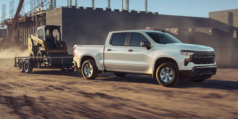 A white Chevrolet Silverado towing a trailer with a yellow skid steer loader across a dusty construction site.